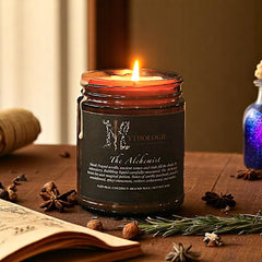 An amber candle with a black label, titled 'The Alchemist', burning on a table with lab equipment and old books in the background, creating a cozy, historical ambiance.
